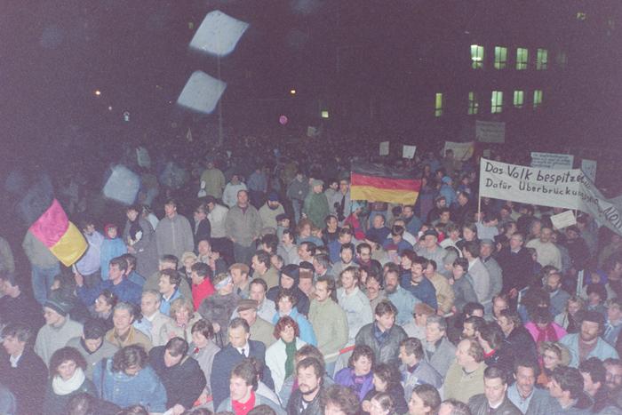 Auf dem Bild sieht man eine große, dicht gedrängte Menschenmenge, die auf das Gelände der Stasi-Zentrale stürmen. Einige Davon halten Transparente oder Deutschlandfahnen in die Höhe.